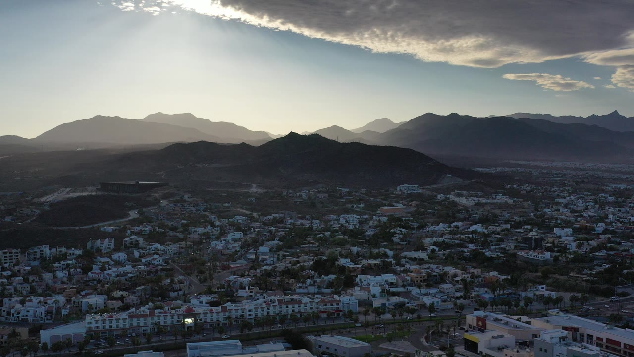 Wide aerial view over Los Cabos, Mexico, with city rooftops in shadow, dark hills in the middle distance, layered mountains on the horizon, and no visible people.
