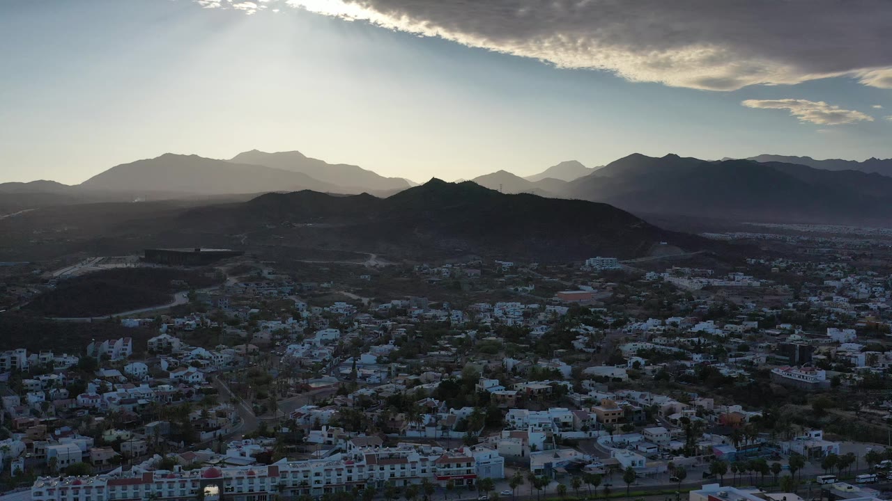Aerial frame over Los Cabos, Mexico, showing the town in shadow, a rounded mountain ridge, layered peaks in haze, bright sun near the cloud edge, and no visible people.