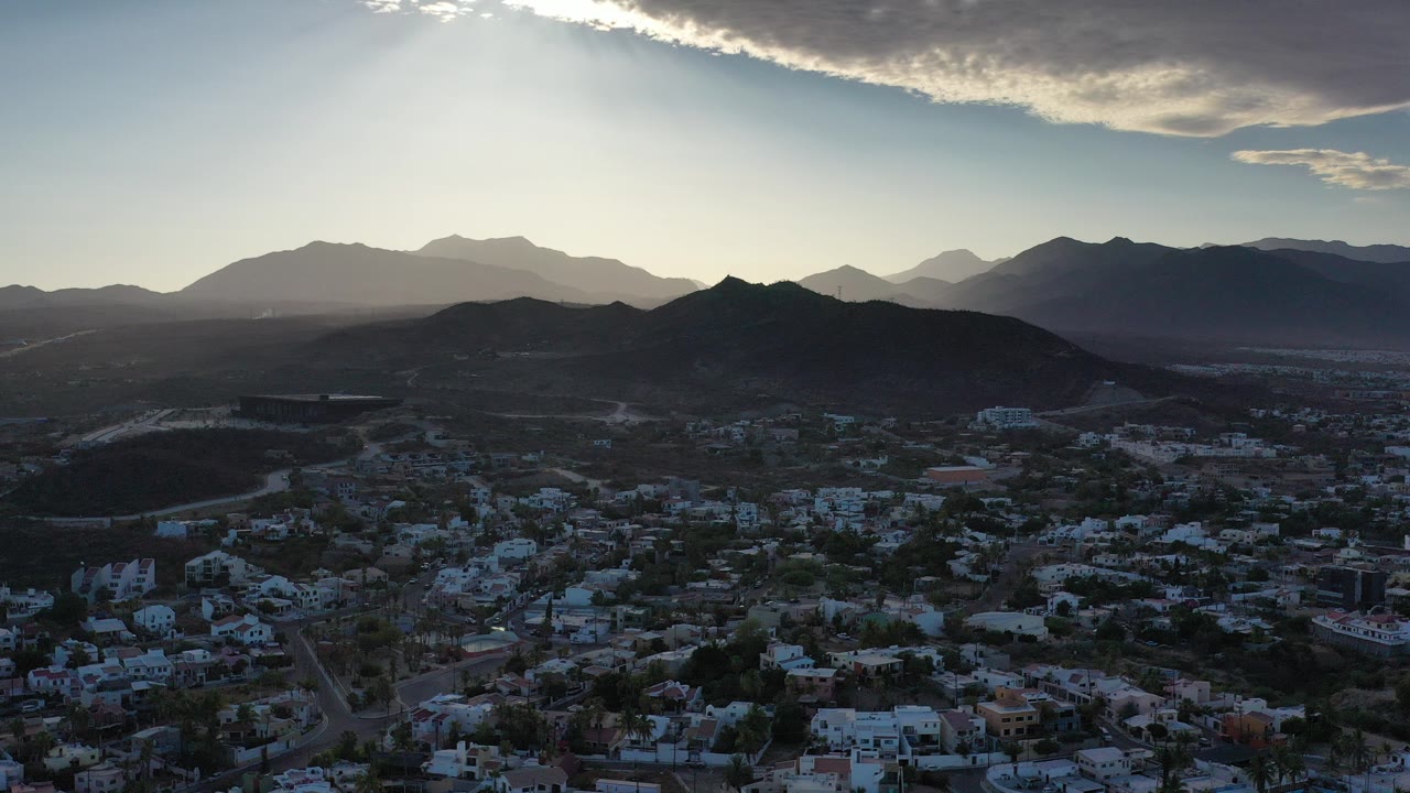 Aerial view over houses, roads, hills, and distant mountains under bright backlight.