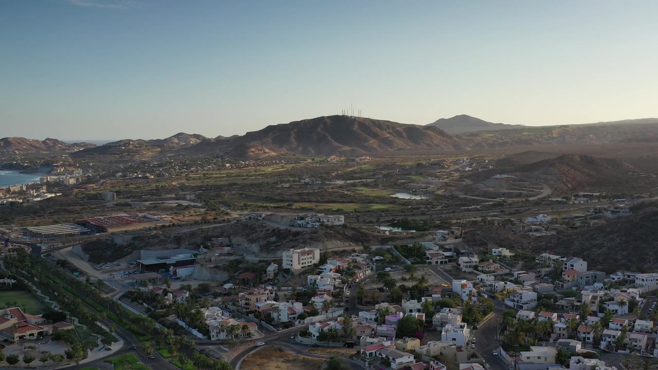 Wide aerial frame over Los Cabos, Mexico, with low-rise homes in the foreground, dry hills across the middle distance, blue water at left, and no visible people.