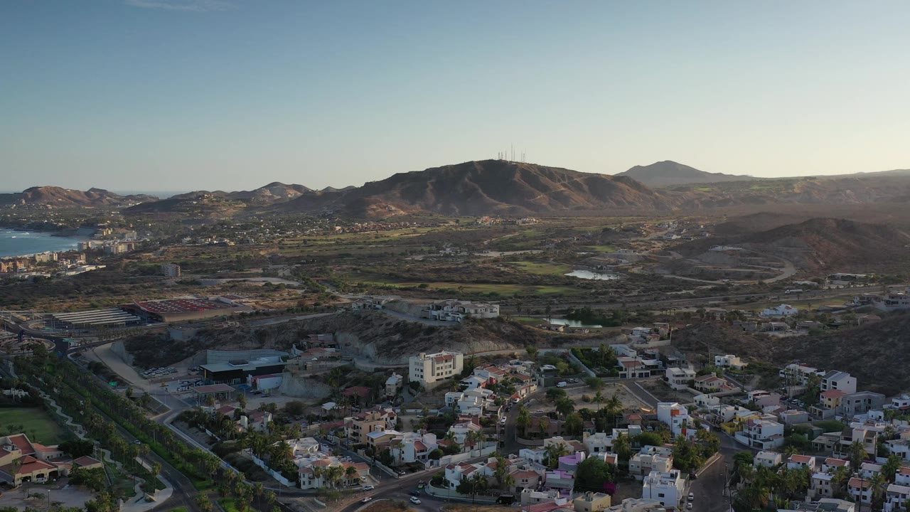 Los Cabos aerial frame with residential blocks at the bottom, a bay or coastal water area on the left, arid hills through the center, and no visible people.
