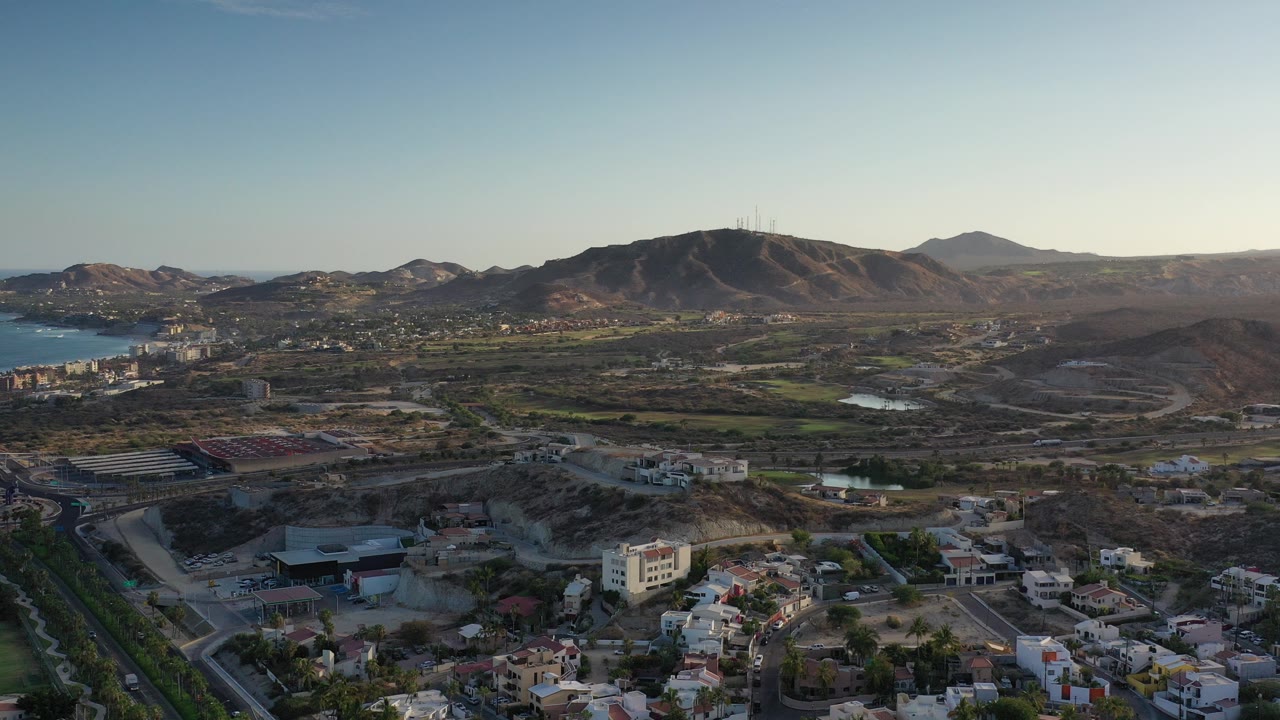 Wide opening aerial over Los Cabos, Mexico, with homes and roads in the foreground, blue water at left, rugged mountains across the horizon, and no visible people.