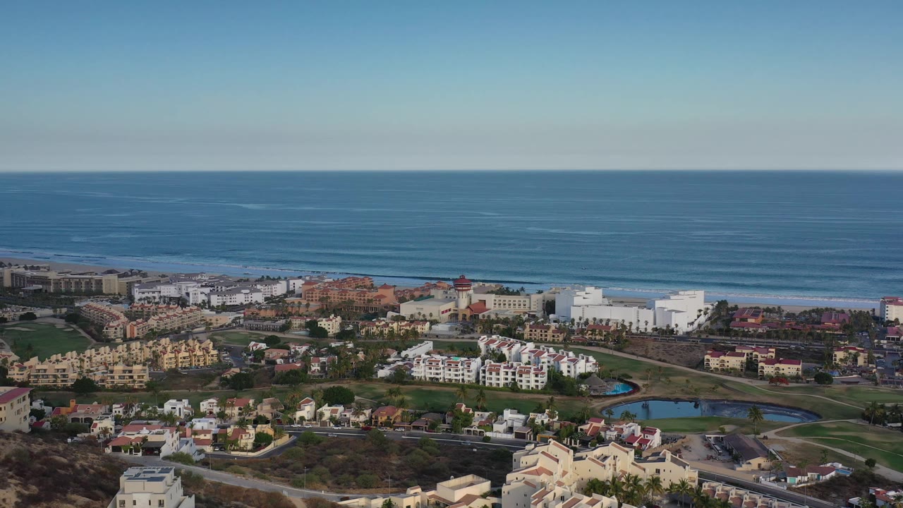 Los Cabos appears from an elevated angle with the ocean across the top of frame, resort-area buildings and homes in the center, and small ponds near the lower edge, with no visible people.