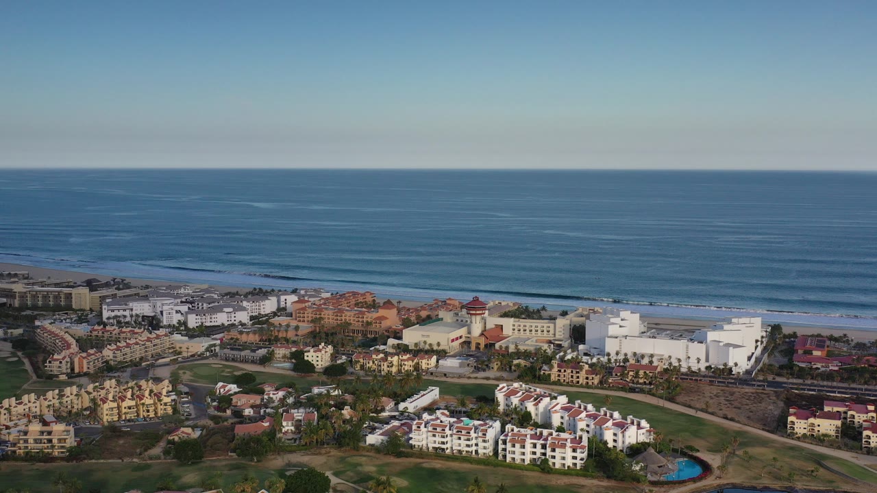 Opening aerial frame over Los Cabos, Mexico, with white and terracotta buildings below, a sandy beach beyond them, blue ocean to the horizon, and no visible people.