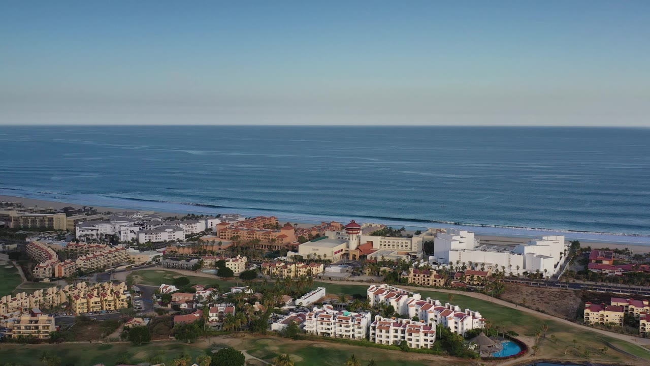 Aerial frame in Los Cabos, Mexico, showing a broad blue ocean horizon above beachfront blocks, villas, roads, and green landscaping, with no visible people.