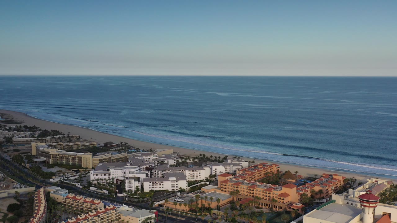 Opening aerial view over Los Cabos, Mexico coastline, with blue ocean, a pale beach, roadside palms, low-rise buildings, and no visible people.
