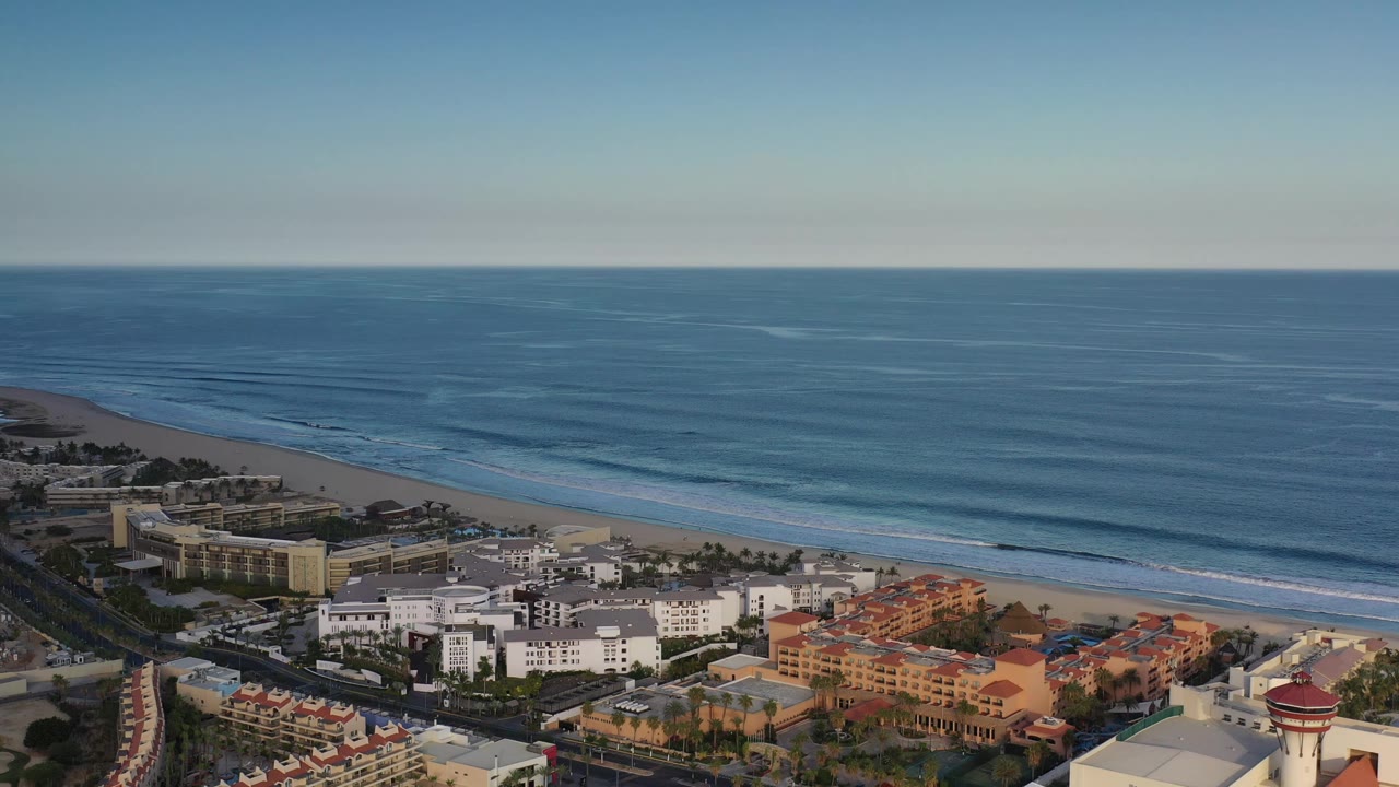 Aerial frame over Los Cabos, Mexico showing the ocean horizon, rolling surf, beachfront buildings, a road lined with palms, and no visible people.