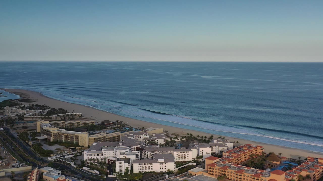 High aerial frame in Los Cabos, Mexico with the beach curving along the left side, waterfront buildings below, long surf lines, and no visible people.