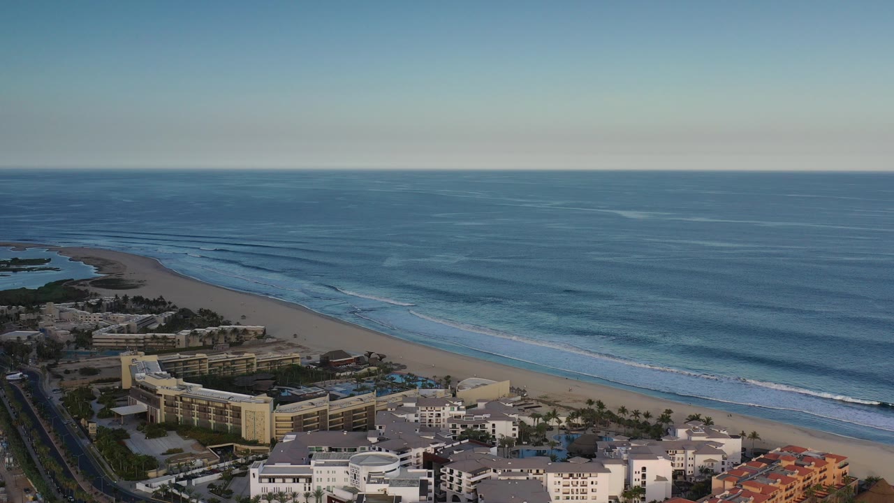Opening aerial frame over Los Cabos, Mexico, showing beachfront buildings, a road, sandy shoreline, blue ocean, white surf bands, and no visible people.