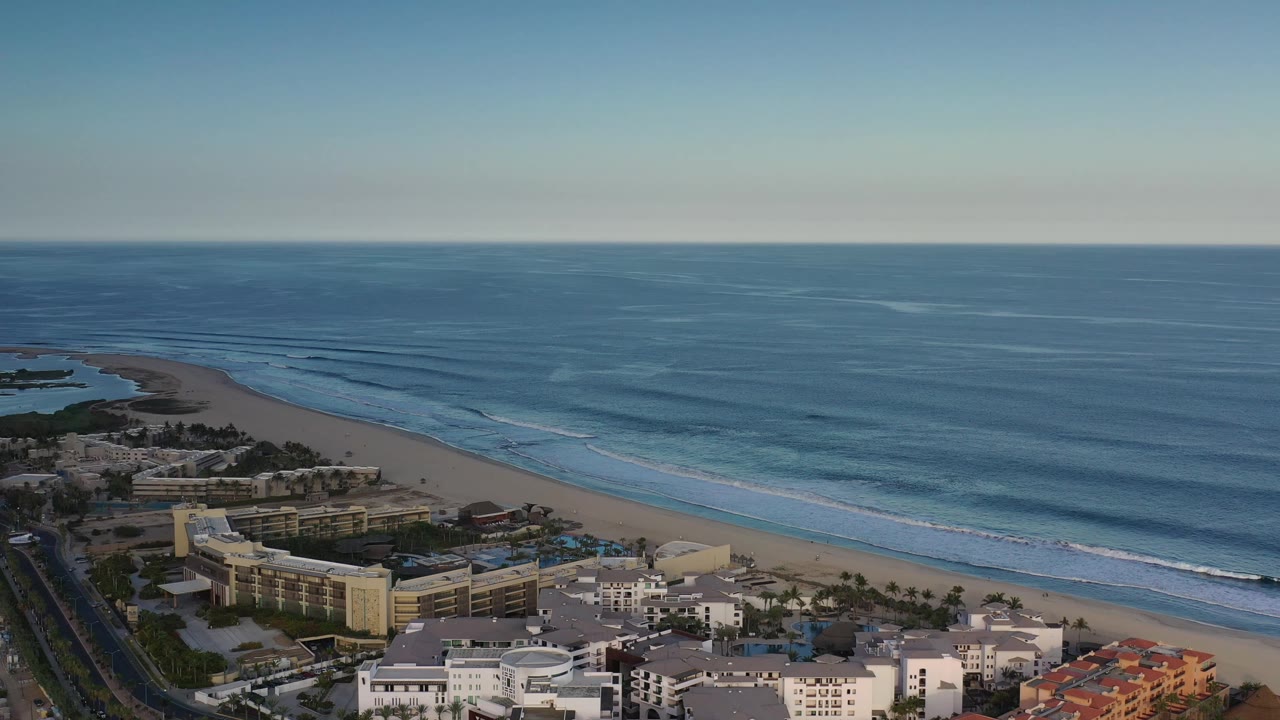 A wide aerial view in Los Cabos, Mexico, with hotel-like buildings low in frame, a long sand strip, calm blue water, white wave lines, and no visible people.