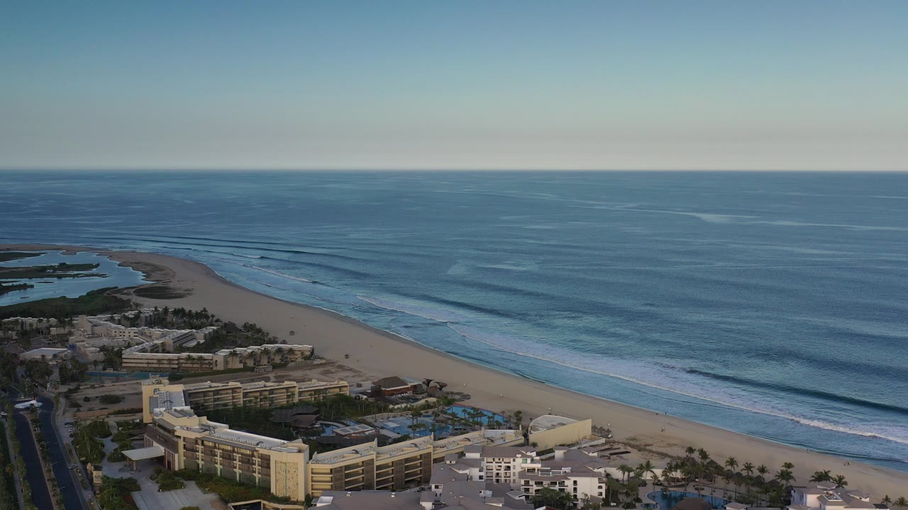 Aerial view over Los Cabos, Mexico with blue ocean, parallel surf lines, sandy beach, beachfront buildings, pools, palms, and no visible people.