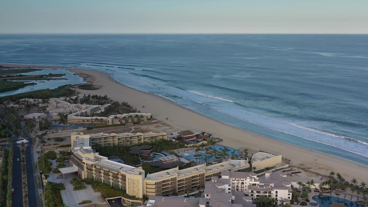 Los Cabos aerial view holding over low buildings and bright pools, with the beach curving toward lagoon water on the left and no visible people.