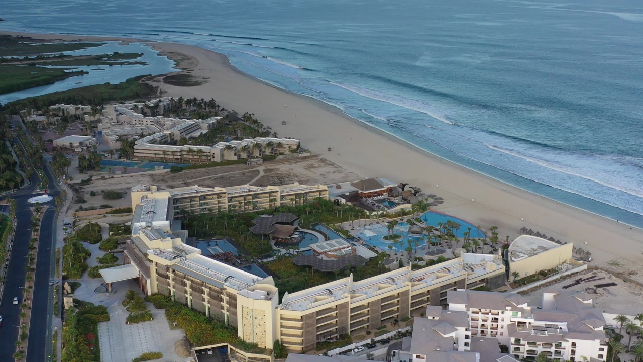 High aerial frame over a Los Cabos beachfront resort with pale hotel buildings, pools, palms, sand, ocean surf, and no visible people.