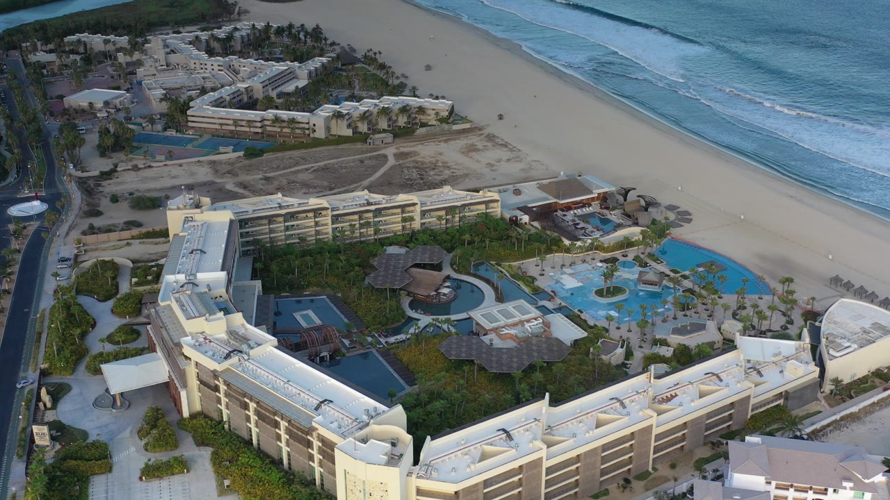 High aerial view over a Los Cabos beach resort with cream buildings in the foreground, blue pools and palm landscaping below, and no visible people.