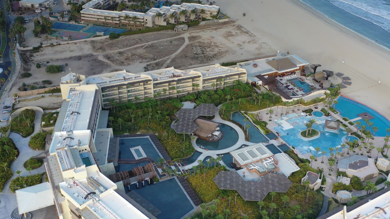 Overhead view in Los Cabos, Mexico showing a resort complex with blue pools, palm landscaping, beach sand, and ocean surf along the top edge, with no visible people.