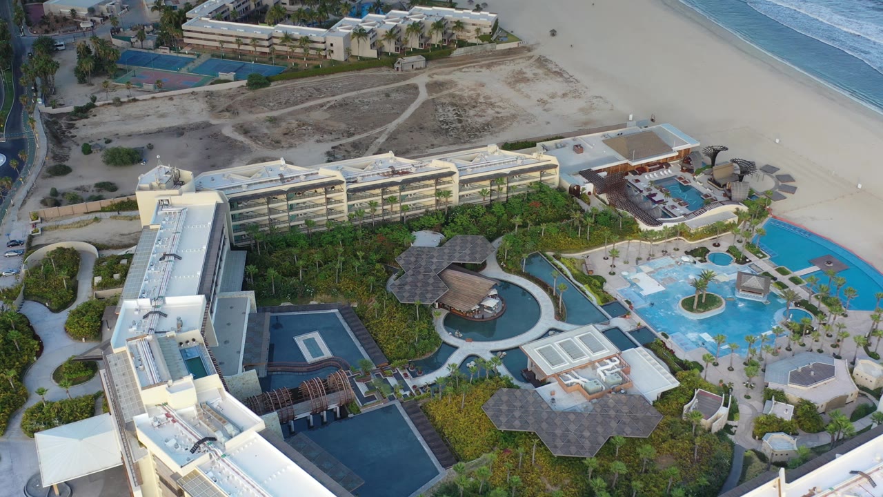 Aerial frame of the Los Cabos resort from above, with a long modern building, dark rooftop areas, curving pool features, sandy beach, and no visible people.