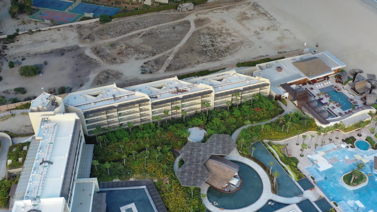 Aerial frame over a Los Cabos resort-style complex with a tall white building at left, blue pools at lower right, landscaped gardens in the center, and no visible people.