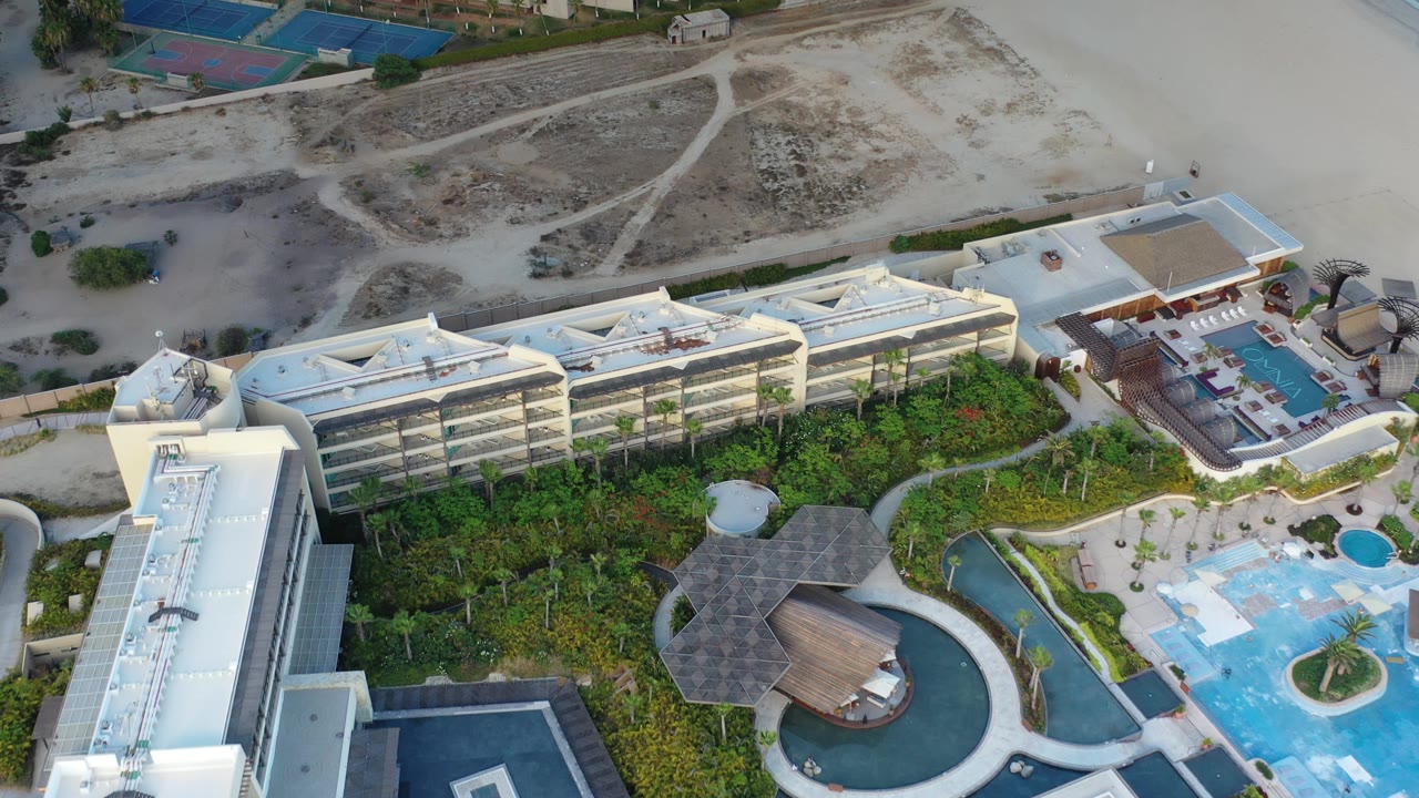 A second aerial moment in Los Cabos shows the long main building across the frame, green garden beds below it, swimming pools to the right, and no visible people.