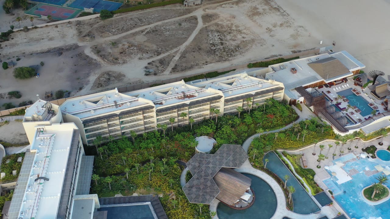 Drone-like overhead view of resort buildings, blue pools, garden paths, and sandy terrain.