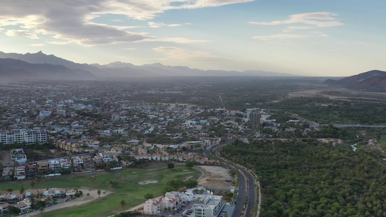 Opening aerial frame over Los Cabos, Mexico, showing a curving highway beside low-rise buildings, green tree cover, grass, hazy mountains, and no visible people.