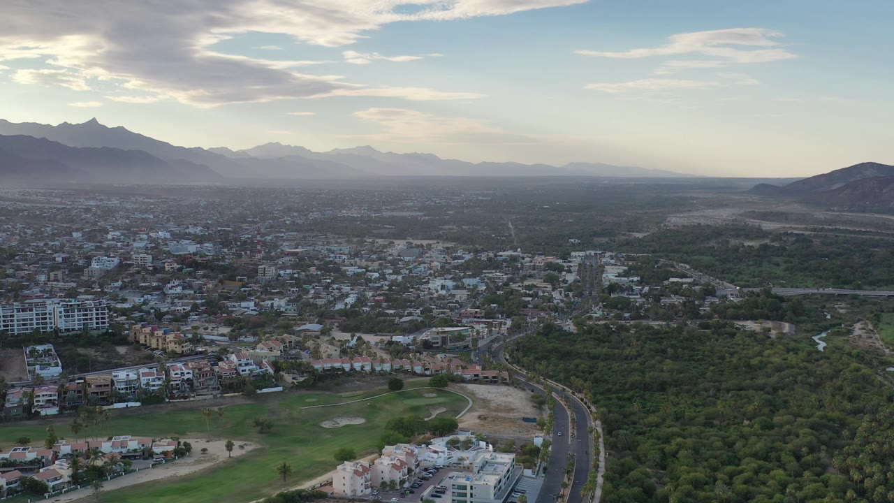 Second aerial frame over Los Cabos, Mexico, with the highway lower in frame, a dense green area through the center, low buildings at left, mountains in the distance, and no visible people.