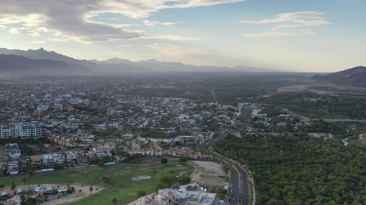 Aerial view over Los Cabos, Mexico with low-rise buildings in the foreground, a curved road at right, green open land through the middle distance, hazy mountains on the horizon, and no visible people.