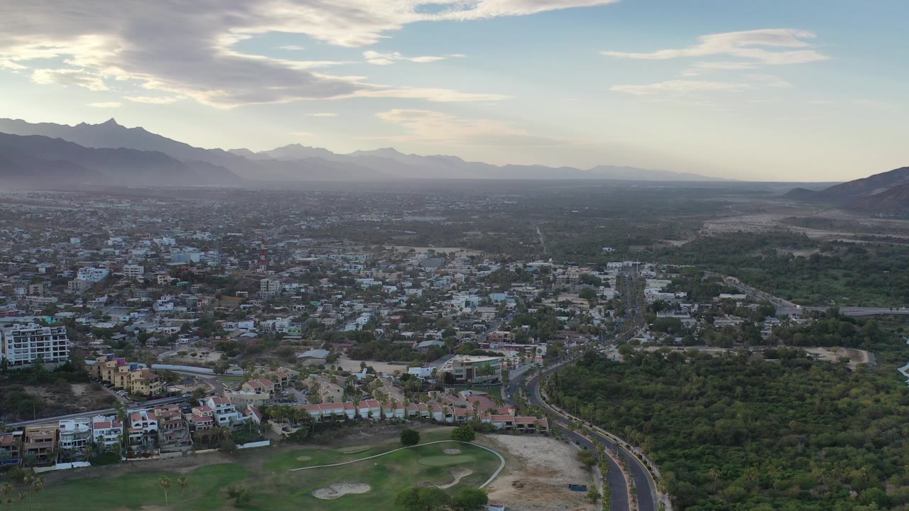 Opening aerial frame over Los Cabos, Mexico, with low-rise buildings, green vegetation, a curving road, distant mountains, pale sky, and no visible people.