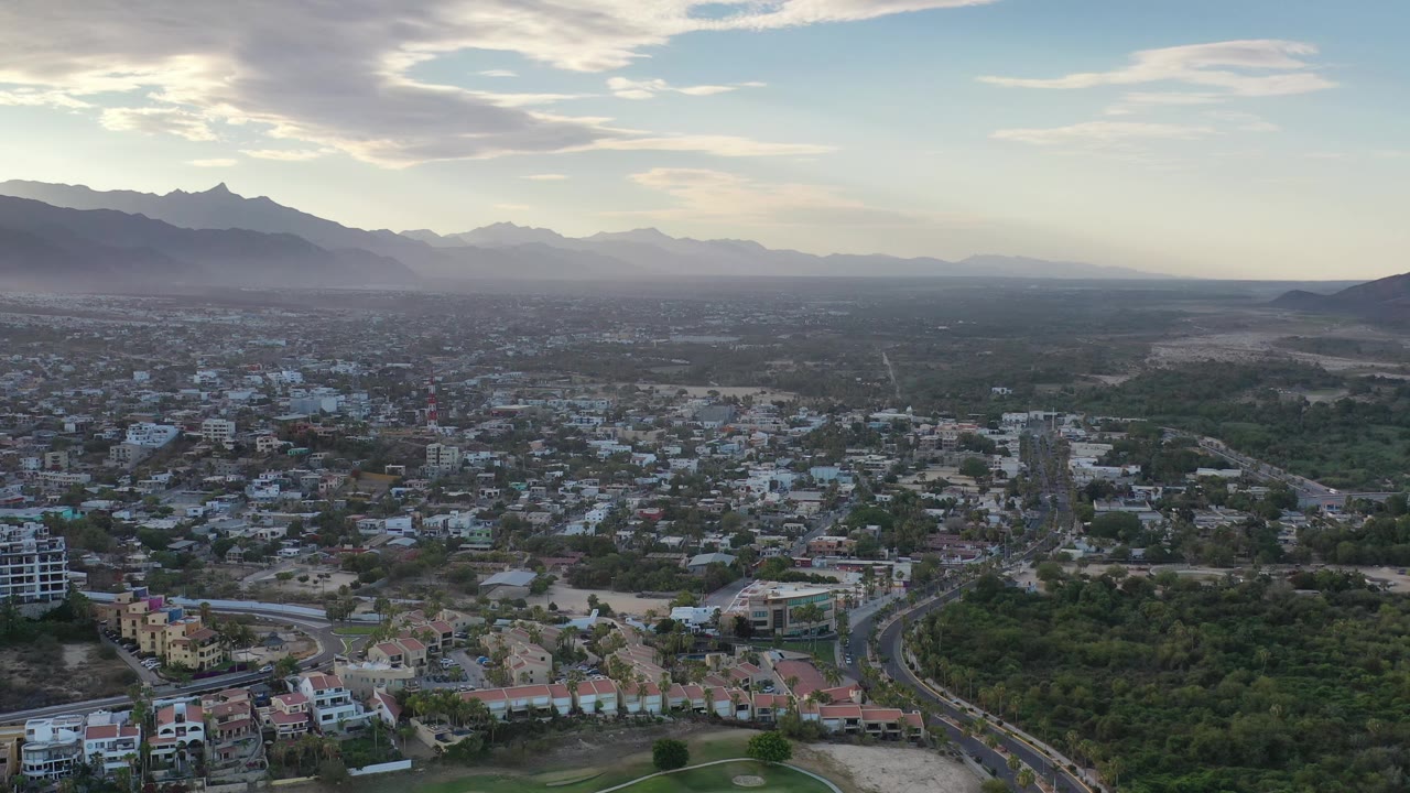 Wide drone-style view of Los Cabos with residential blocks and roads in the lower frame, a dark green corridor, distant mountains, and no visible people.