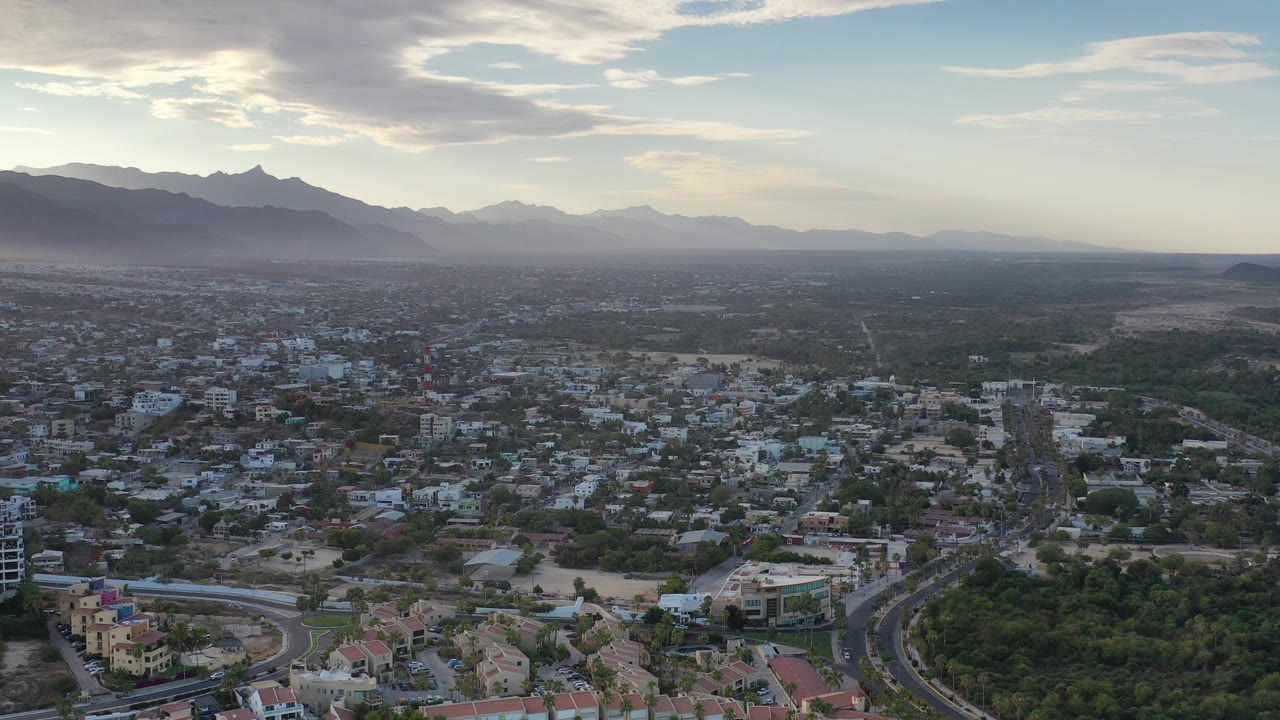 Opening aerial view over Los Cabos, Mexico, showing low buildings, curved roads, dry lots, green areas, hazy mountains, and no visible people.