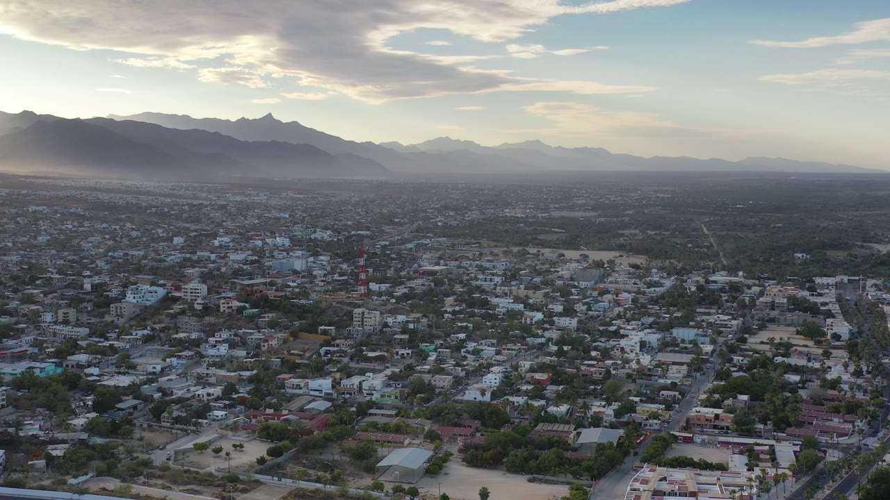 Wide aerial view over a Los Cabos, Mexico residential area with low-rise homes, streets, distant mountains, hazy light, and no visible people.