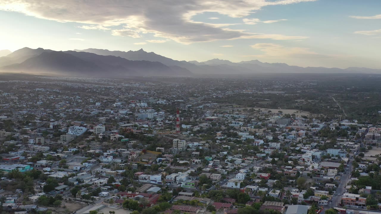 Los Cabos aerial frame with residential-looking rooftops, a straight road through the lower center, layered mountain ridges in haze, bright cloud cover, and no visible people.
