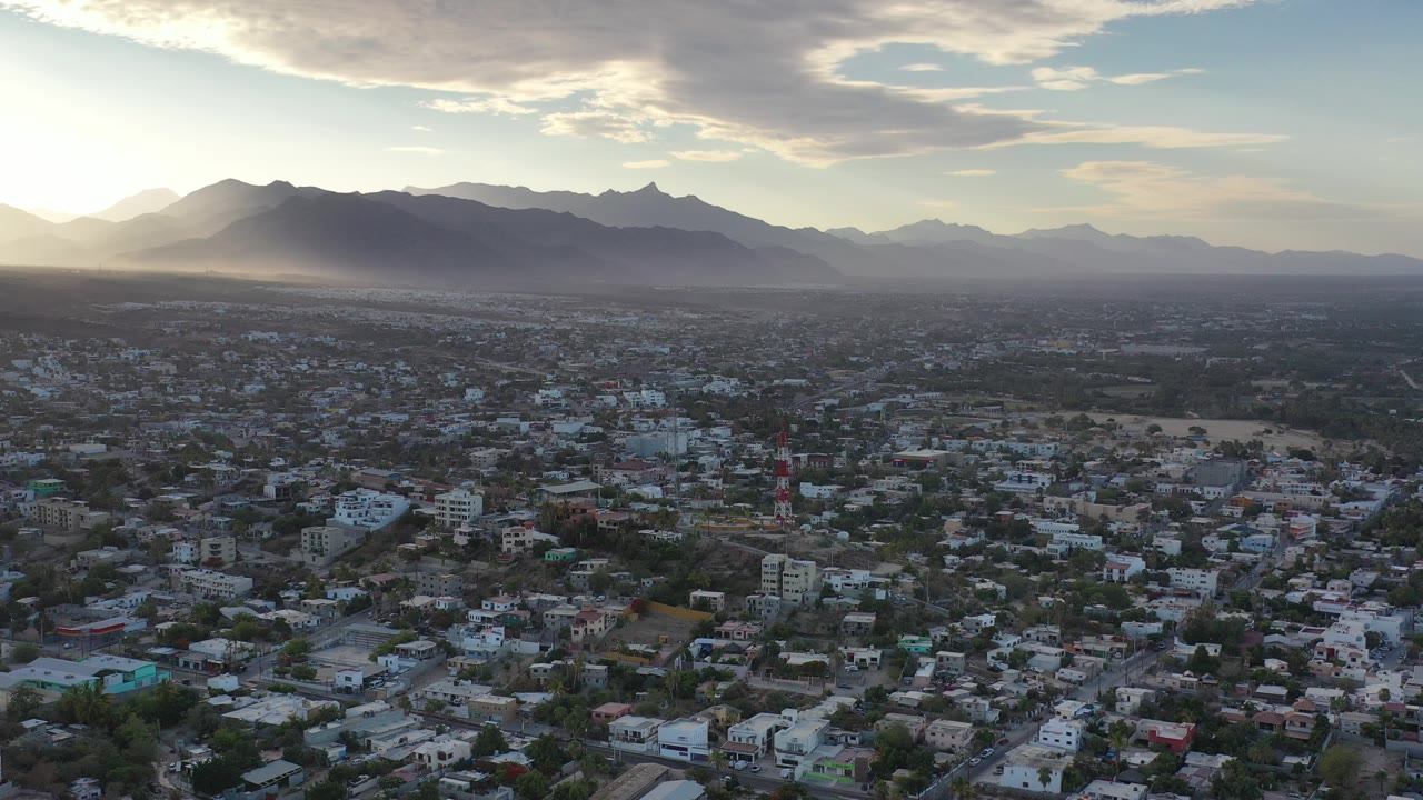 Wide aerial view over Los Cabos, Mexico, with dense city blocks below, dark mountain ridges on the horizon, low sun at left, layered clouds above, and no visible people.