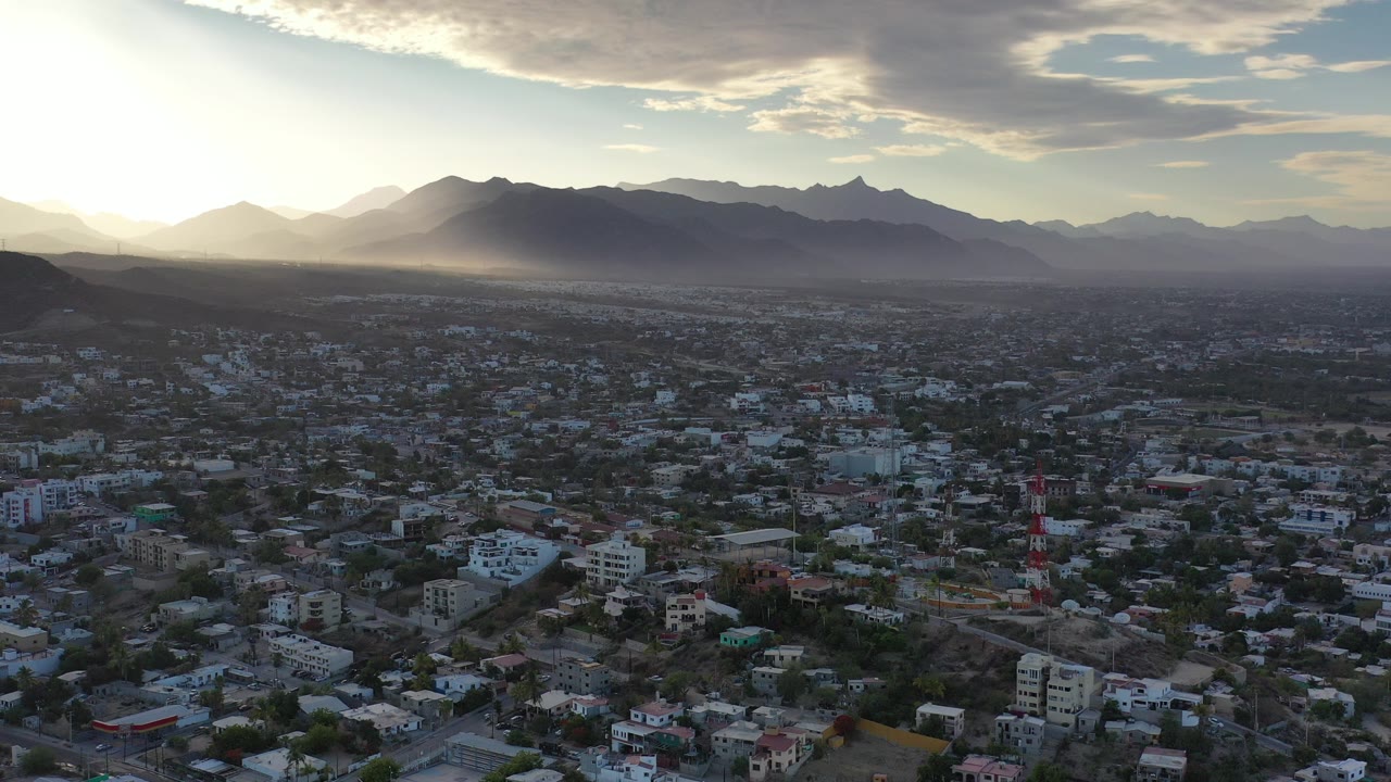 Aerial view of city buildings below hazy mountains at low sun.