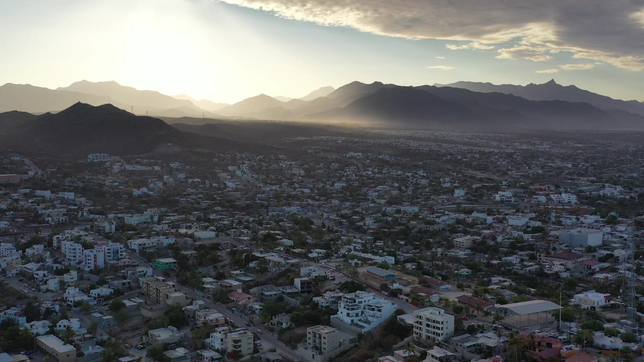 Opening frame of Los Cabos, Mexico from above, with city buildings below, layered mountains on the horizon, strong sun at upper left, and no visible people.