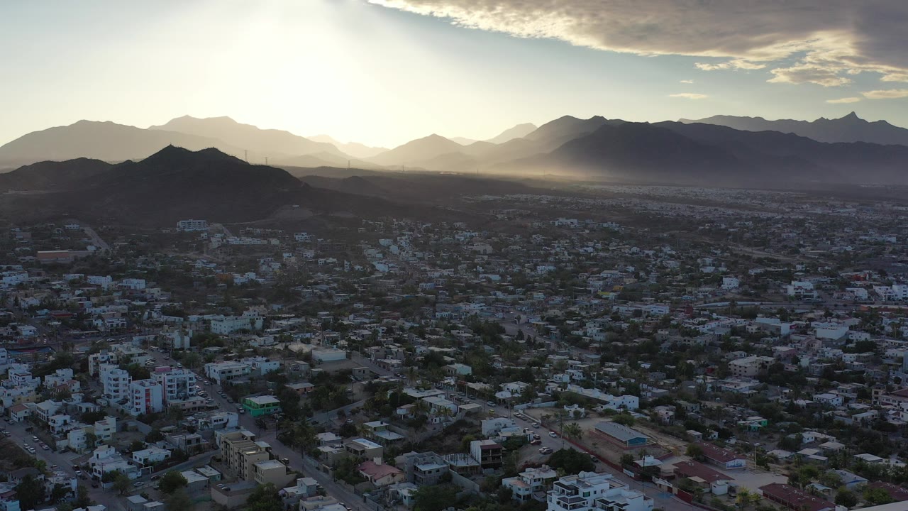 Aerial view over Los Cabos, Mexico with low-rise city blocks below layered mountain ridges, bright sun haze near the horizon, and no visible people.