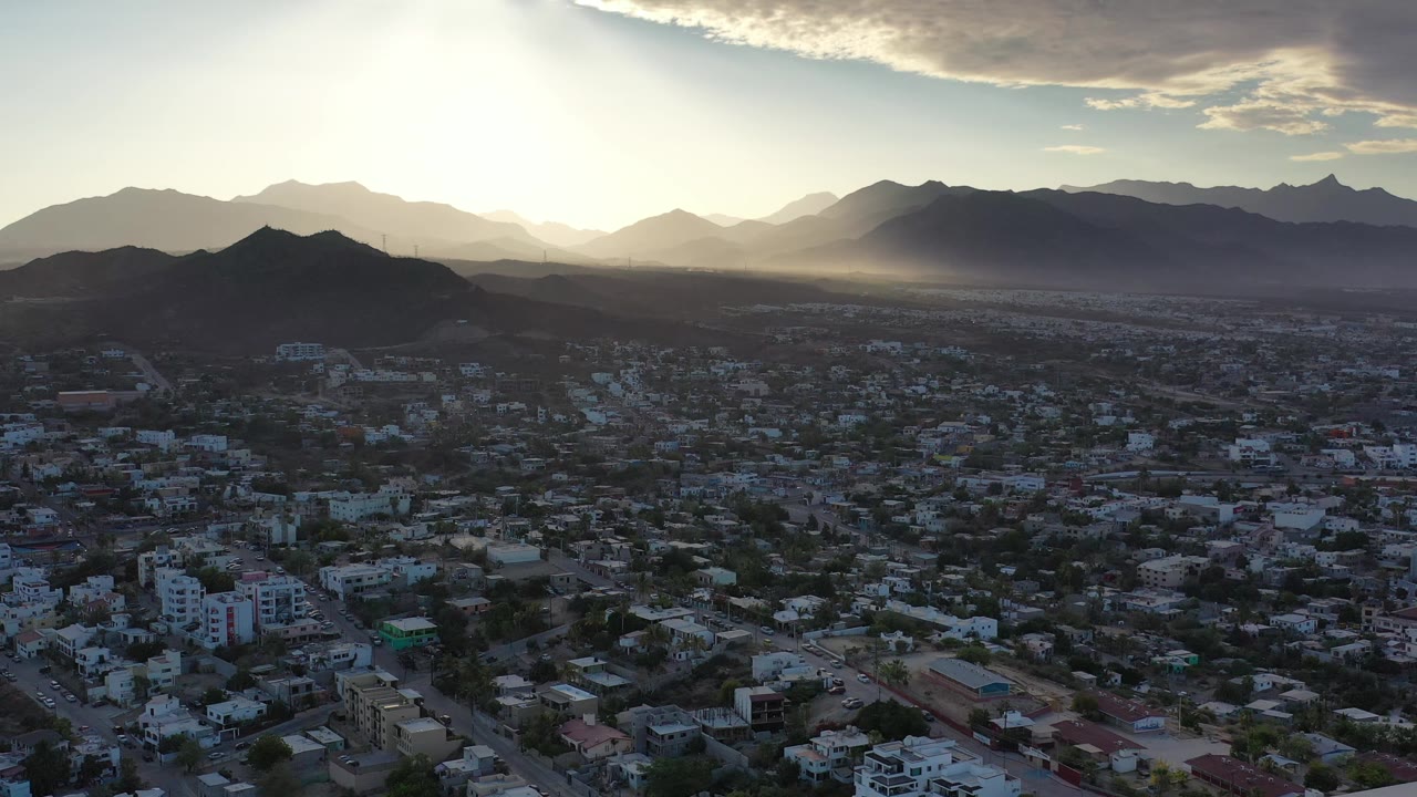 Los Cabos, Mexico appears from above with dark foreground neighborhoods, a rounded ridge at left, distant peaks, strong sun glare, and no visible people.