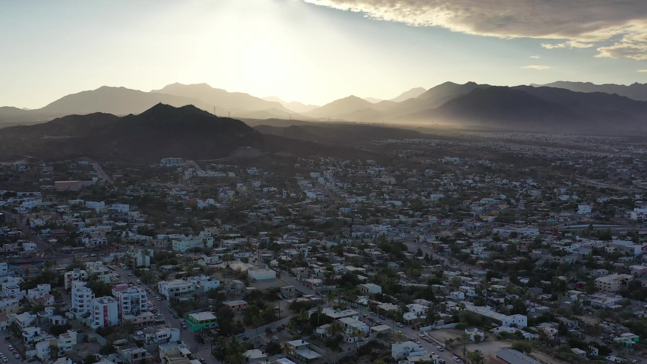 Aerial frame over Los Cabos, Mexico, showing darker foreground rooftops, layered mountain silhouettes, a bright sunlit haze behind the peaks, and no visible people.