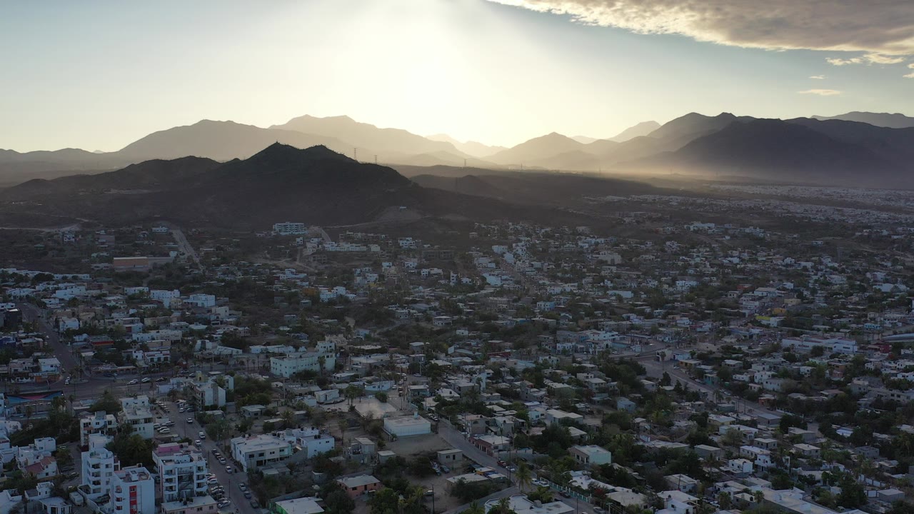 Aerial view over Los Cabos with compact town blocks in the foreground, dark hills across the middle, bright low sun behind distant mountains, and no visible people.