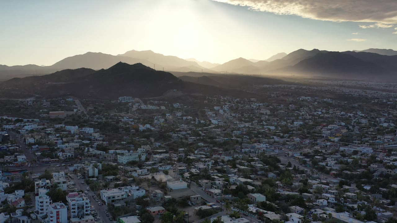 Los Cabos appears from above with residential and commercial-looking buildings below, a dark hill ridge near center, pale mountains beyond, and no visible people.