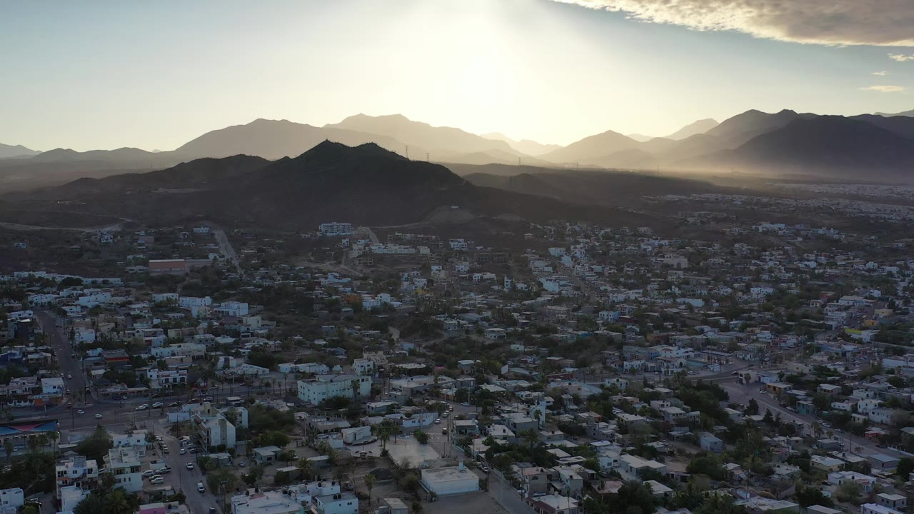 Los Cabos, Mexico appears from a high aerial angle with shadowed neighborhoods, a dark hill across the center, layered mountains, sun flare, and no visible people.