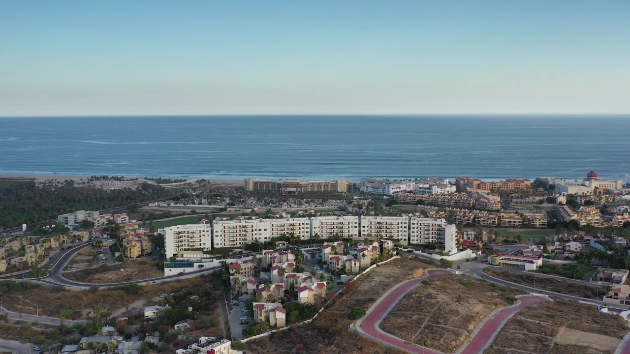 Opening frame of Los Cabos, Mexico from above, showing apartment blocks, small homes, curving roads, dry ground, and a broad ocean horizon with no visible people.
