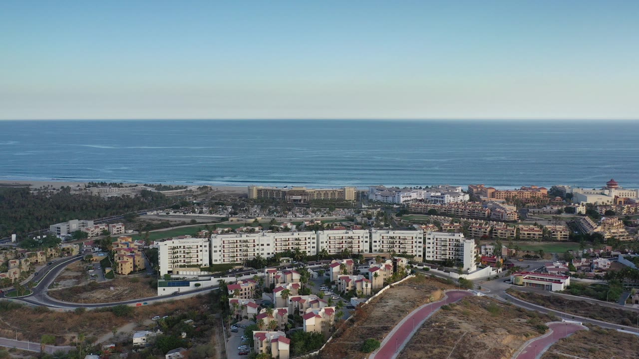Opening aerial frame over Los Cabos, Mexico shows white condo buildings, dry lots, curving roads, beach vegetation, and a blue ocean horizon with no visible people.