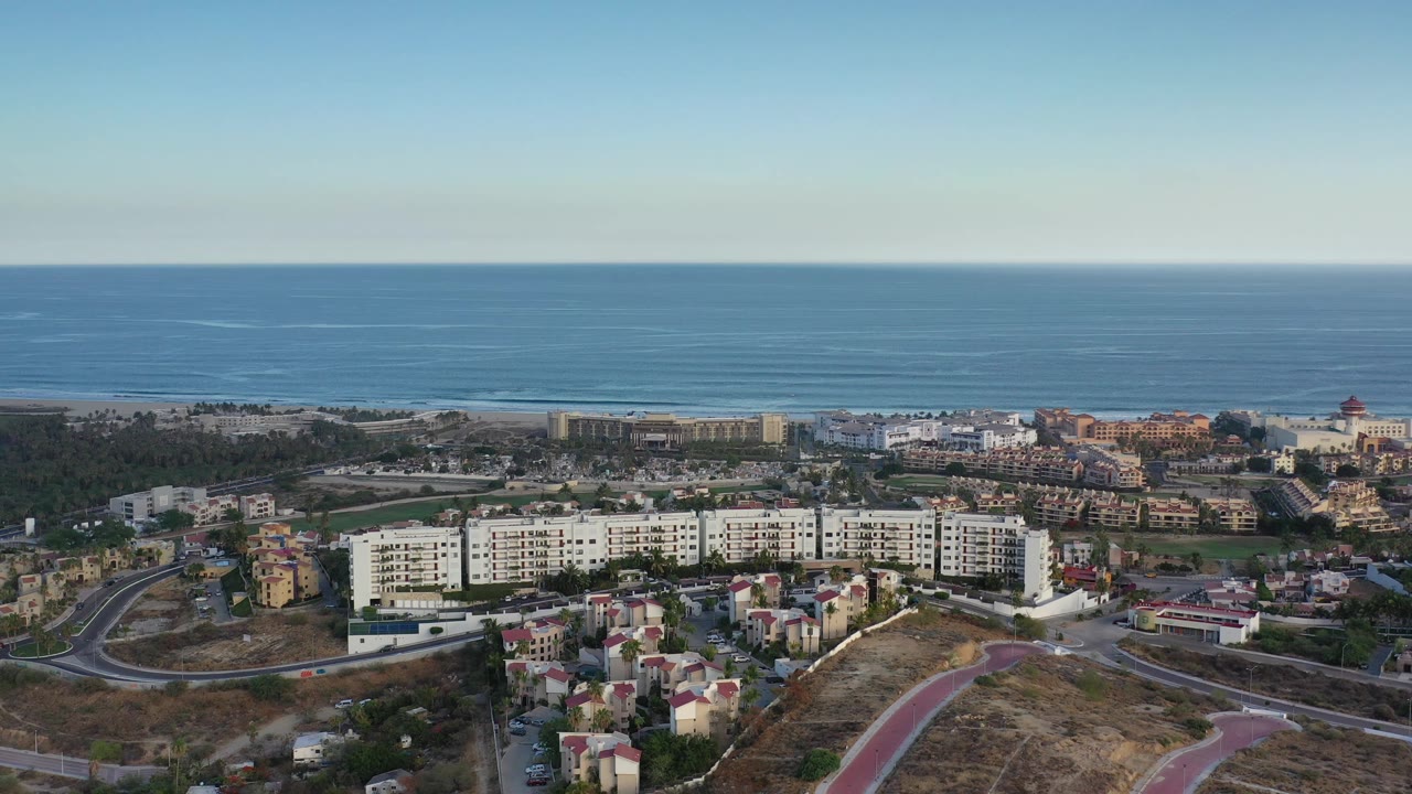Early aerial moment in Los Cabos, Mexico keeps the white apartment blocks centered, with smaller homes below, a dry slope at right, and the ocean horizon across the top with no visible people.