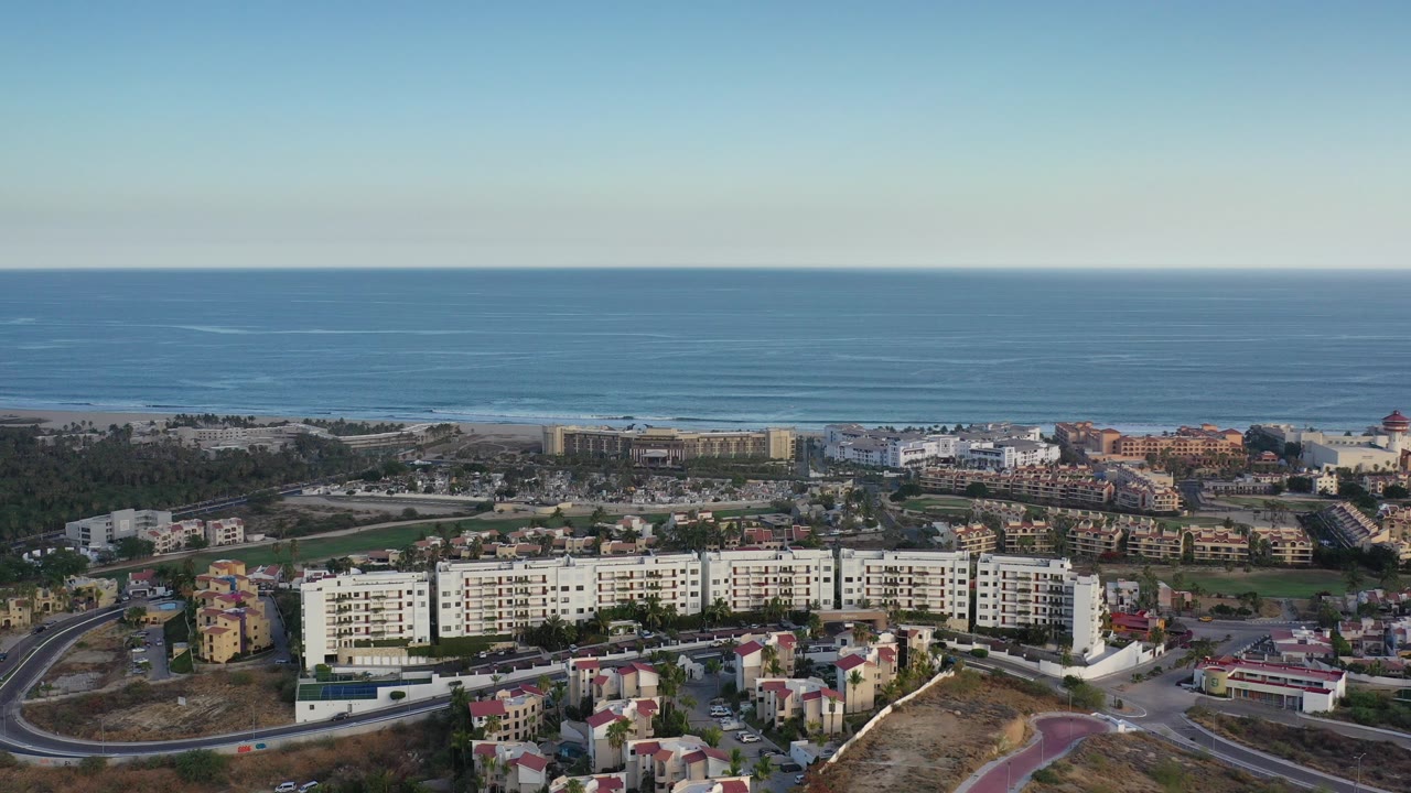 Opening aerial frame over Los Cabos with white apartment blocks, roads, palms, beachfront buildings, blue ocean, pale sky, and no visible people.
