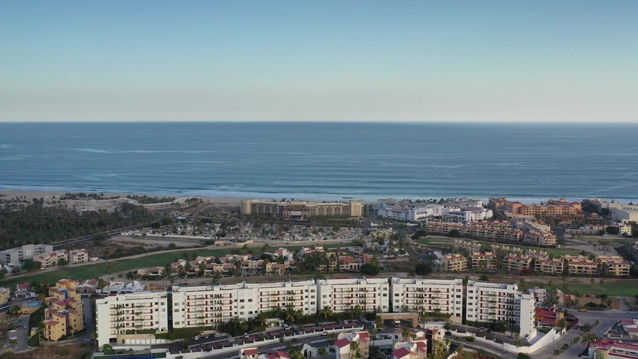 Aerial view in Los Cabos, Mexico, showing a coastal neighborhood, white multi-story buildings at the bottom edge, green trees, and a wide blue ocean backdrop with no visible people.