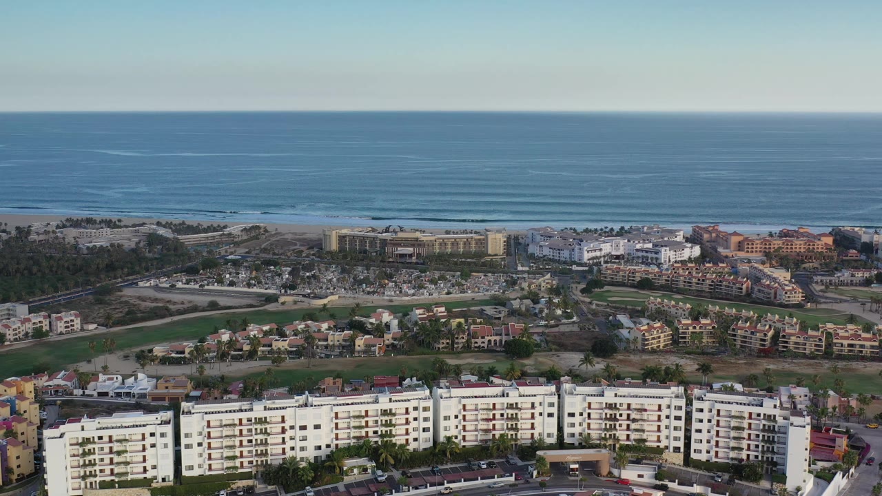 Aerial view in Los Cabos, Mexico, showing blue ocean across the top, beach development in the center, and white multi-story buildings along the lower edge with no visible people.
