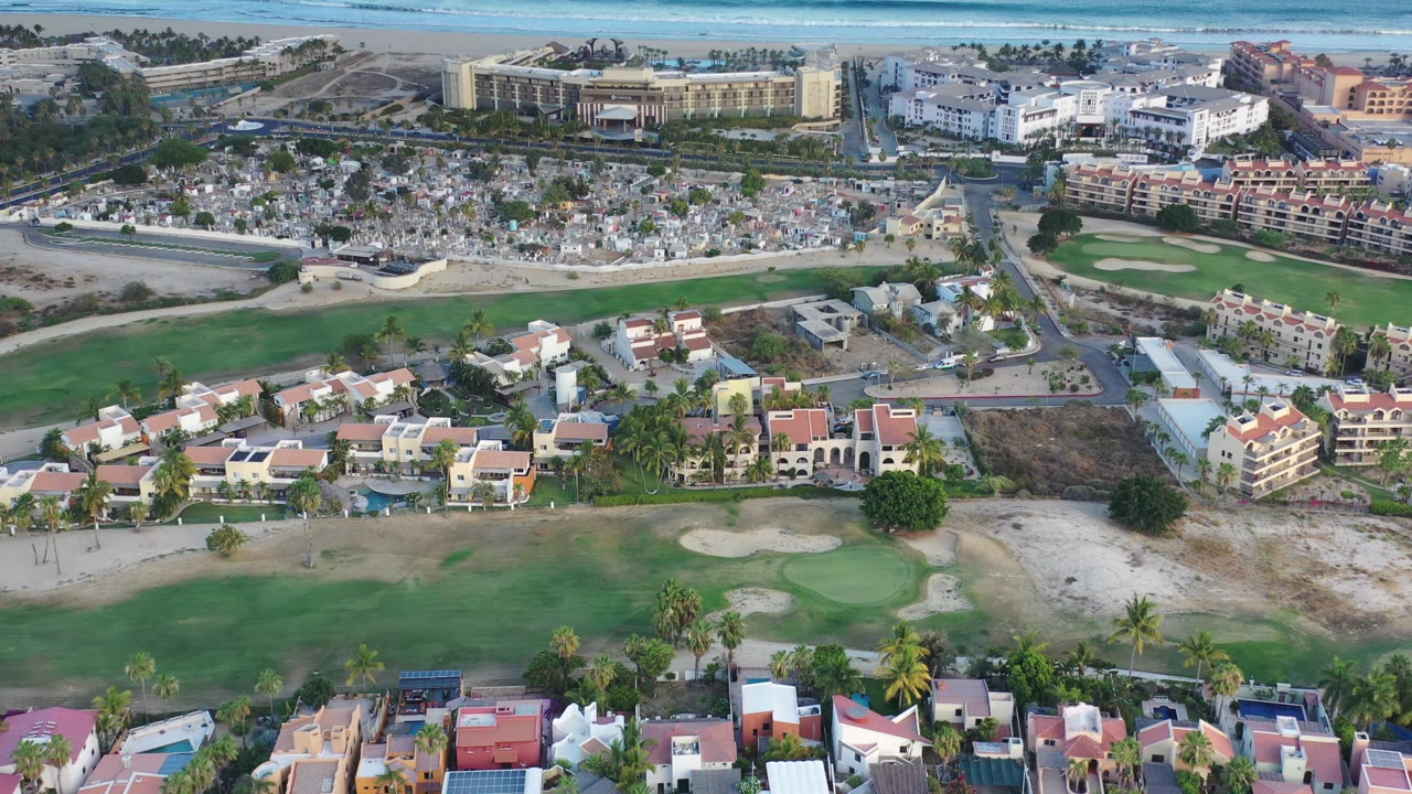 Los Cabos aerial view holds over a fairway and sand traps, with houses, a road junction, a cemetery, low-rise buildings, and no visible people.