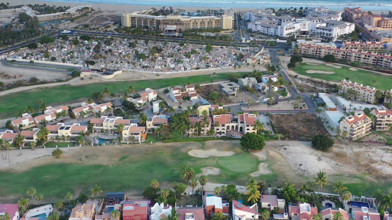 Drone-height view of homes bordering a golf course with cemetery and water behind.