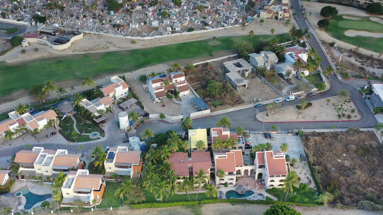 High aerial frame over Los Cabos luxury houses with red tile roofs, private pools, a curved street, green course grass, dry lots, and no visible people.