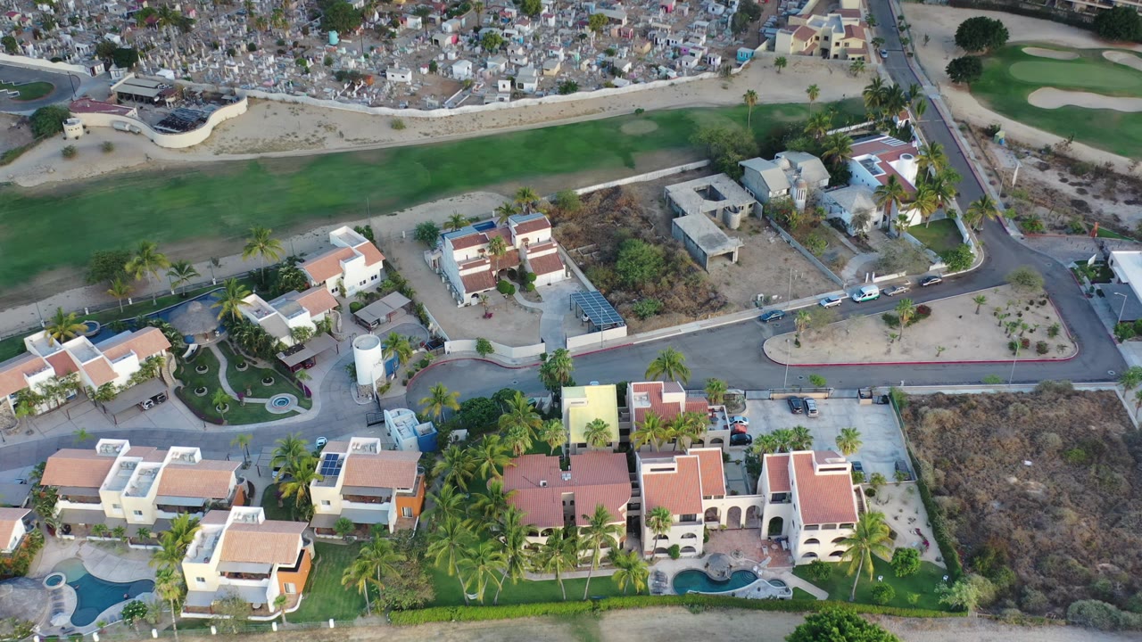 Aerial view in Los Cabos showing modern villas, blue swimming pools, palm-lined yards, a road intersection, dry open parcels, and no visible people.